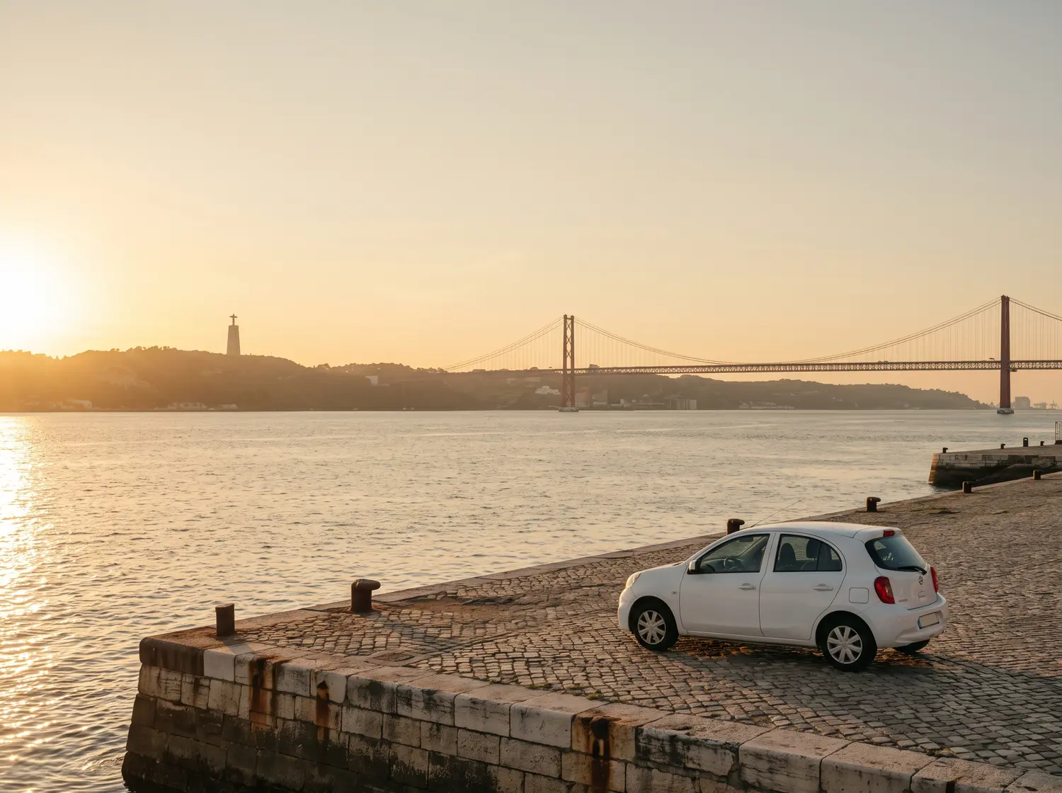 A white small hatchback parked on a cobbled quay at sunset, with a long red suspension bridge across the water beyond